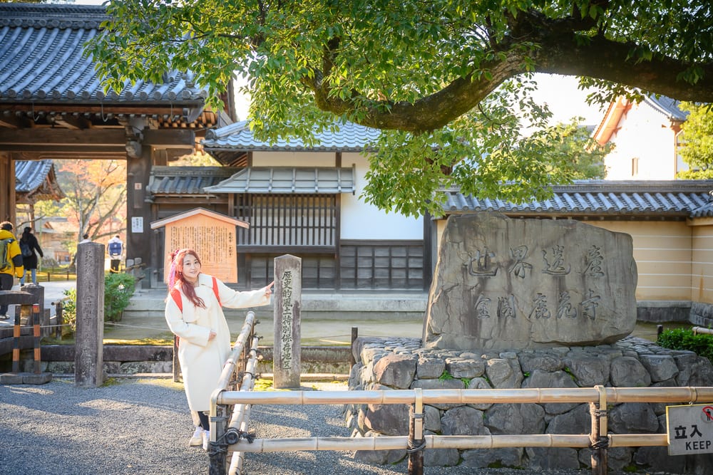 日本京都金閣寺完整指南|池畔倒影與庭園步道 10 日本京都金閣寺|寺廟、石碑、木造建築、女子、樹木,女子身穿白色長大衣站在石碑旁,指向刻有文字的石柱,前方有竹製圍欄,背景為傳統日式瓦屋頂與木門。