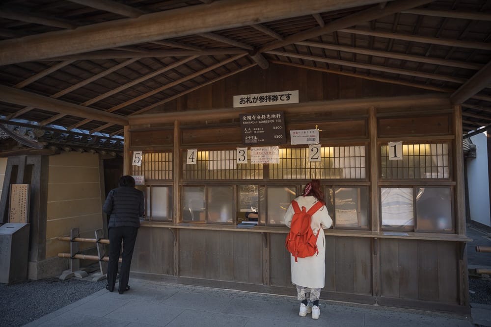 日本京都金閣寺完整指南|池畔倒影與庭園步道 13 日本京都金閣寺|木造售票亭、窗戶、告示牌、遊客。兩名遊客分別站在編號窗戶前,木造屋簷下光線均勻,窗上方懸掛日文招牌與票價表。