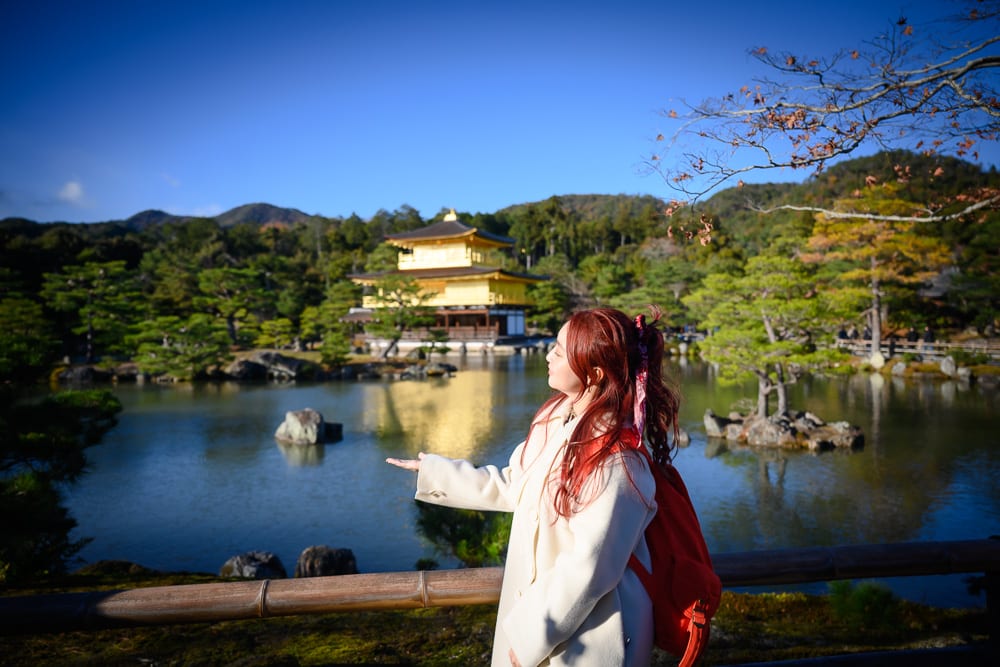 日本京都金閣寺完整指南|池畔倒影與庭園步道 12 日本京都金閣寺|女子、寺廟、池塘、山景、樹木、竹欄杆,紅髮女子背紅背包穿白大衣立於湖畔,單手平舉對向遠方金色寺廟。