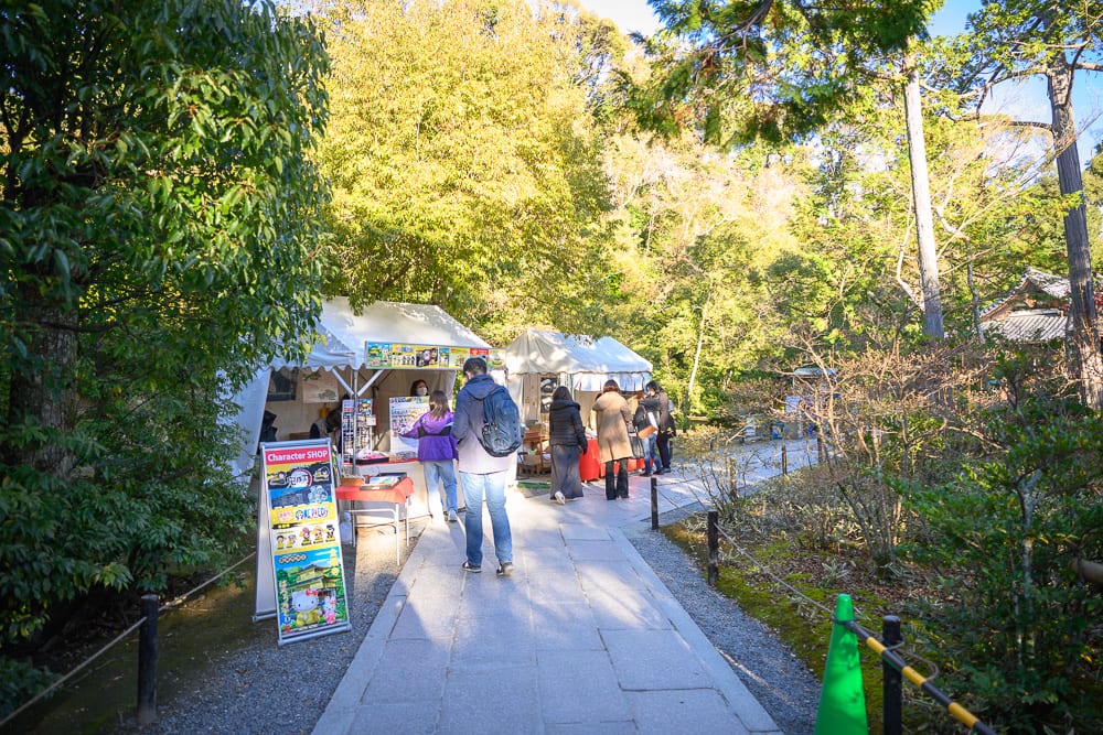 日本京都金閣寺完整指南|池畔倒影與庭園步道 6 日本京都金閣寺|石板路、帳篷、商店、遊客、樹林,戶外步道旁設置白色販賣攤位,行人駐足挑選商品,兩側被茂密綠樹與枝葉包圍。