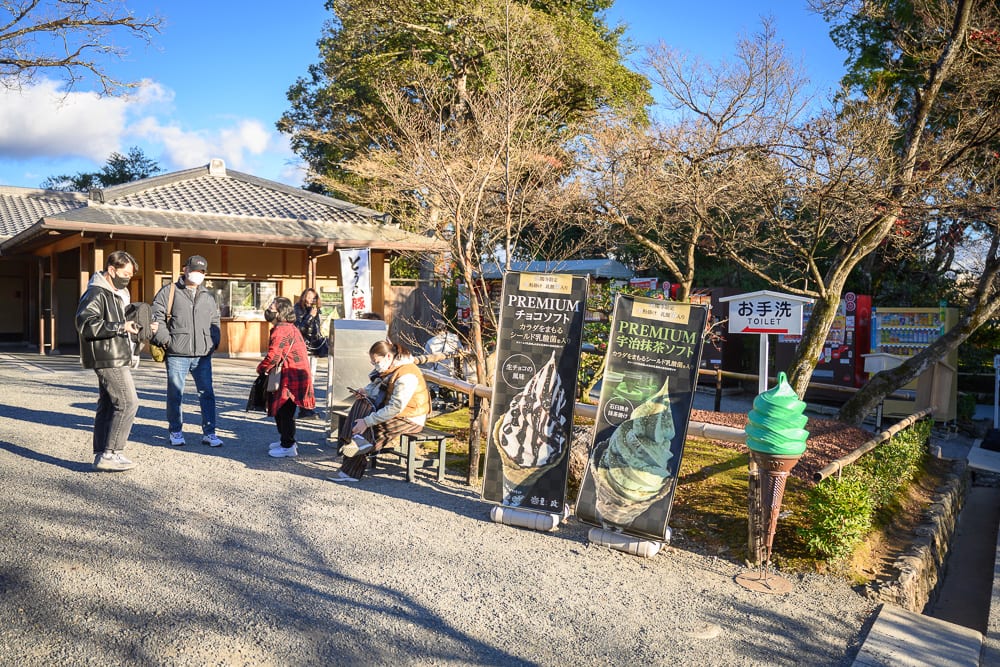 日本京都金閣寺完整指南|池畔倒影與庭園步道 3 日本京都金閣寺|街道,行人,冰淇淋招牌,日式平房,枯樹。戶外廣場上,幾名戴口罩的行人站立與坐著休息,前方豎立著霜淇淋圖案的直立招牌與指標,右側擺放綠色冰淇淋模型,背景為傳統屋瓦建築。