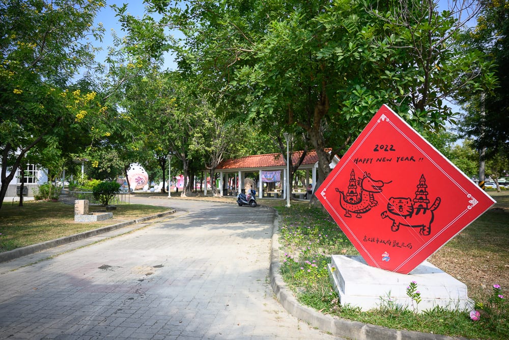 高雄蓮池潭兒童公園｜戶外公園，石磚步道，綠樹，涼亭，菱形新年裝飾牌。紅底看板繪有龍虎圖案與2022字樣，前方草地平坦，背景有白色建築與遠方雕塑。