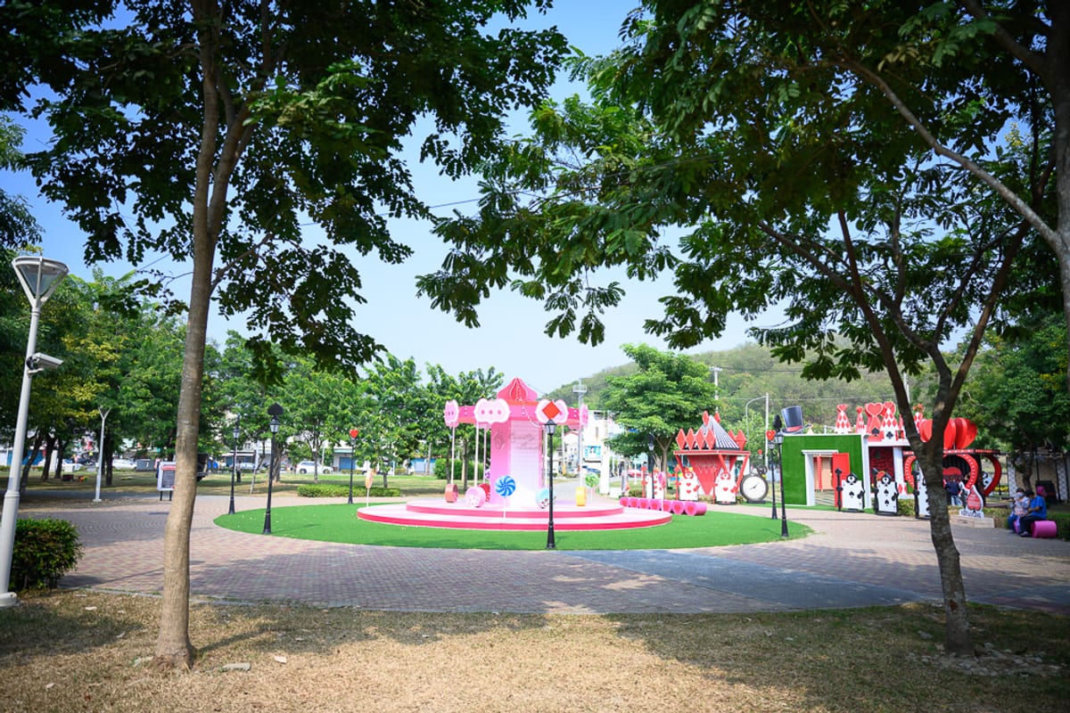 高雄蓮池潭兒童公園｜戶外公園，粉紅旋轉木馬，綠地，樹木，磚造廣場，路燈，遠方綠樹山丘。晴朗天光下，木馬與愛麗絲主題裝置坐落於圓形草皮中央，周圍散布著路燈與鋪面廣場。