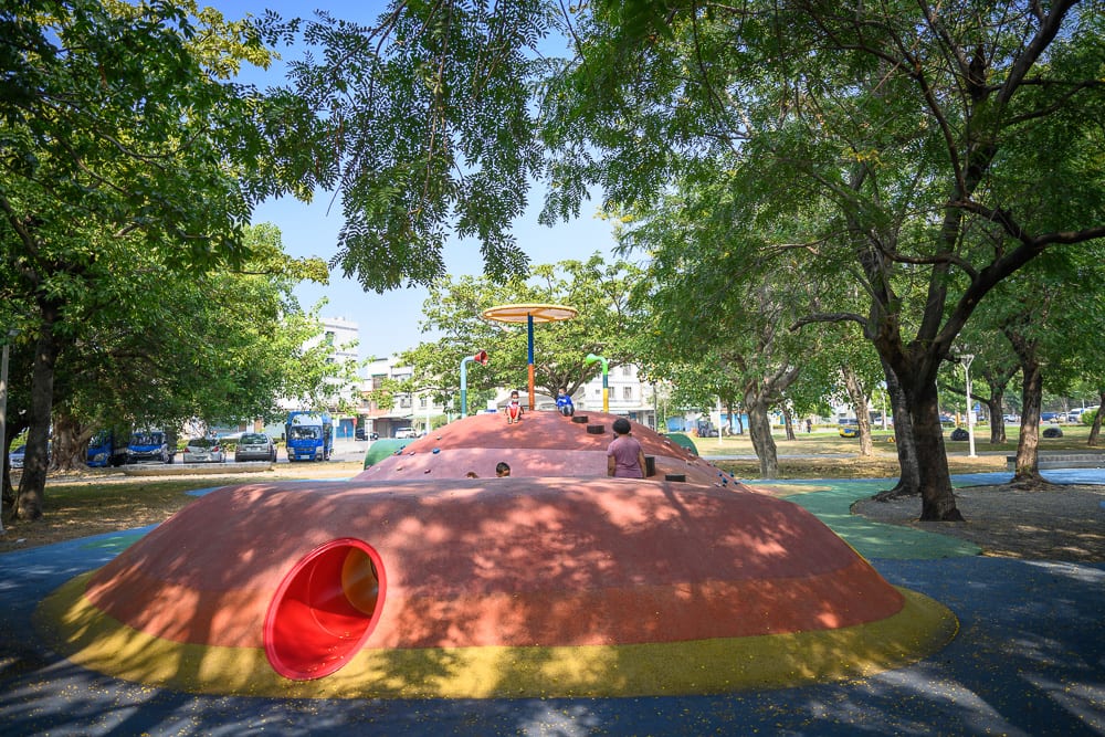 高雄蓮池潭兒童公園｜公園、遊樂場、圓形土丘、攀爬架、隧道、大樹、汽車，綠樹成蔭的戶外空間中，有著紅黃相間的錐形遊戲設施，幾位兒童在土丘上方活動，周邊停放多輛車輛。