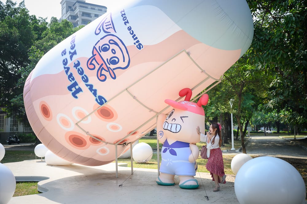 高雄蓮池潭兒童公園｜戶外公園、巨型充氣飲料杯、卡通吉祥物公仔、長裙女子，綠樹環繞草皮，地面散落白色圓球，女子手部觸碰公仔背部，斜向巨大杯身印有珍珠圖案與文字。