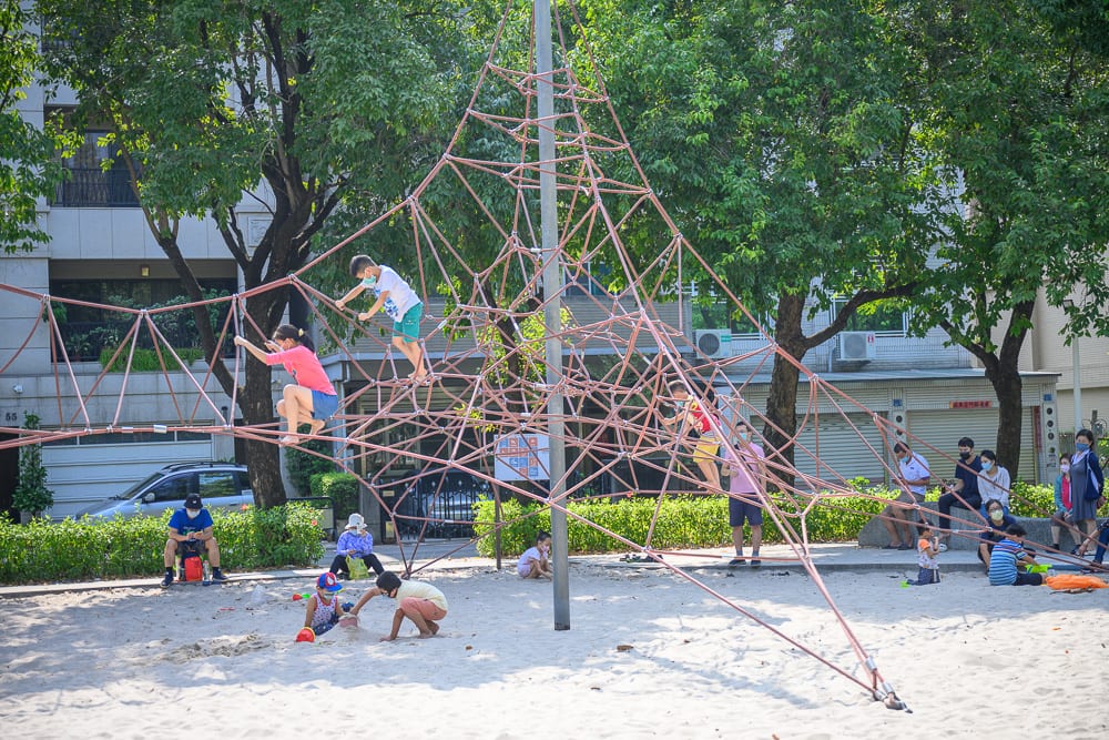 高雄蓮池潭兒童公園｜戶外公園，沙坑，攀爬網，孩童，成人，樹木，建築物。多位孩童在大型錐形繩網上攀爬，周圍沙坑裡有孩童坐地玩沙，周邊座椅有家長坐著看顧，背景為綠樹與住宅建築。