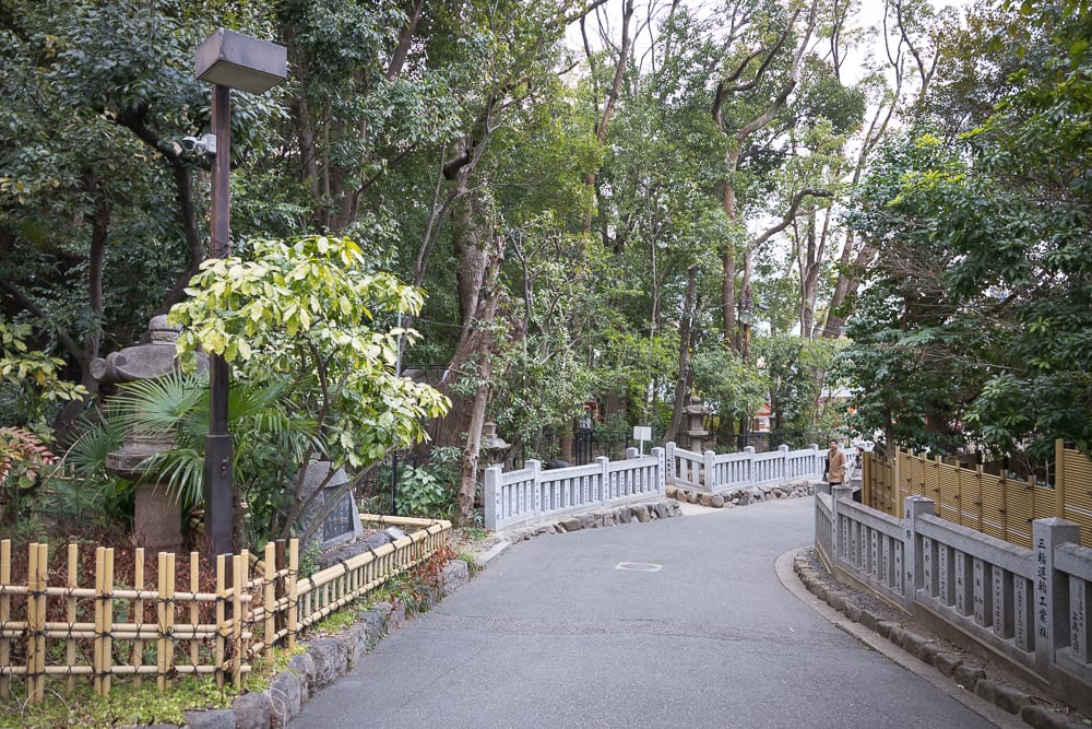 日本神戶-生田神社｜柏油路、石燈籠、路燈桿、竹籬笆、石製欄杆、樹木。彎曲的柏油路穿梭於茂密樹林間，路旁設有低矮竹籬與石造護欄，左側立有高聳路燈與石燈籠，遠處可見石柱與行人身影。