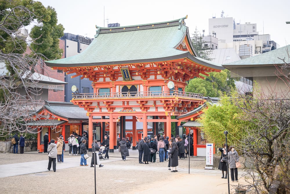 日本神戶-生田神社｜寺廟、人群、街道、建築、樹木，橘紅色雙層木造寺門佇立於廣場中央，綠色斜屋頂層疊，大批遊客穿梭於門廊下與前方石板地，背景夾雜現代高樓與枯枝。