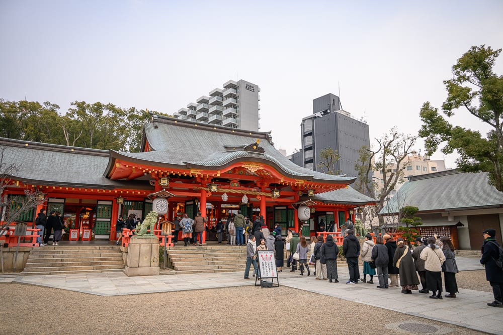 日本神戶-生田神社｜寺廟、階梯、人群、高樓，橘紅色傳統神社建築前鋪滿石子地，參拜民眾排成一列等待進入殿內，後方矗立著現代水泥公寓與灰色大樓。