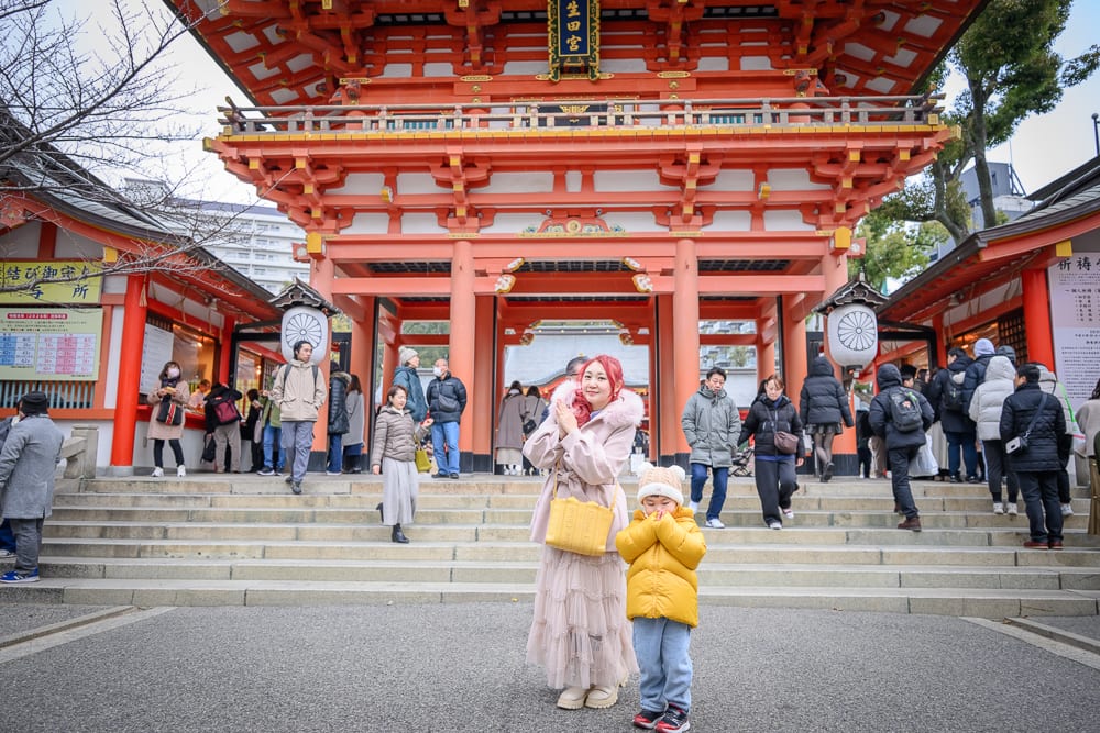日本神戶-生田神社｜寺廟、階梯、人群、冬衣、木造建築。朱紅色的大型寺廟門前，石階上有穿著厚外套的眾多遊客走動。前景站著兩位戴著毛帽與冬衣的女子和幼童，背後是懸掛圓形燈籠的雙層牌樓。