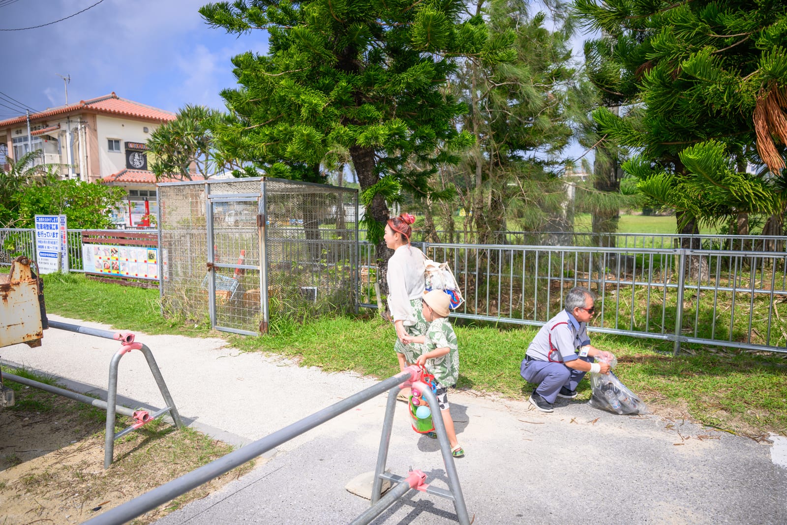 日本沖繩恩納村海濱公園｜戶外道路、鐵絲網欄杆、金屬柵欄、綠樹、矮牆建築、女性、兒童、男性。路面上有金屬阻隔架，草地上有鐵籠與蹲姿男性手提垃圾袋。