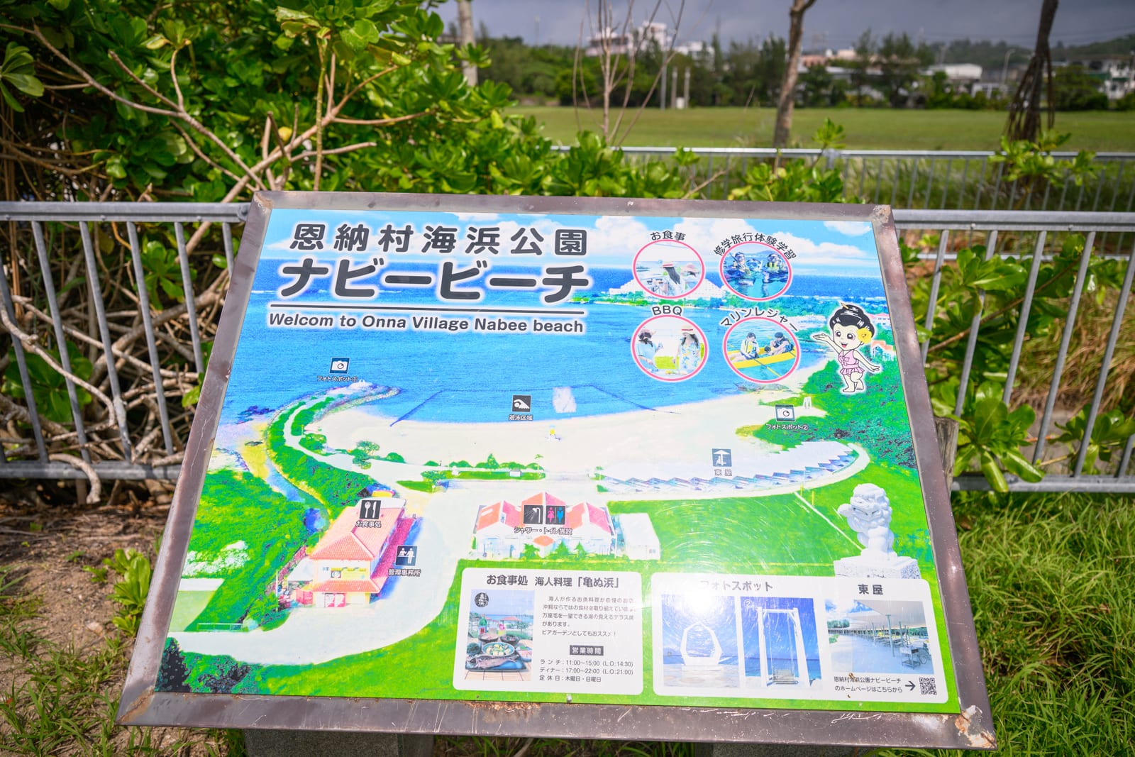 日本沖繩恩納村海濱公園｜告示牌、公園地圖、海灘、鐵欄杆、草地，金屬支架撐起戶外導覽看板，表面印有園區設施配置圖與日文說明，背景為綠色草皮與金屬圍欄。
