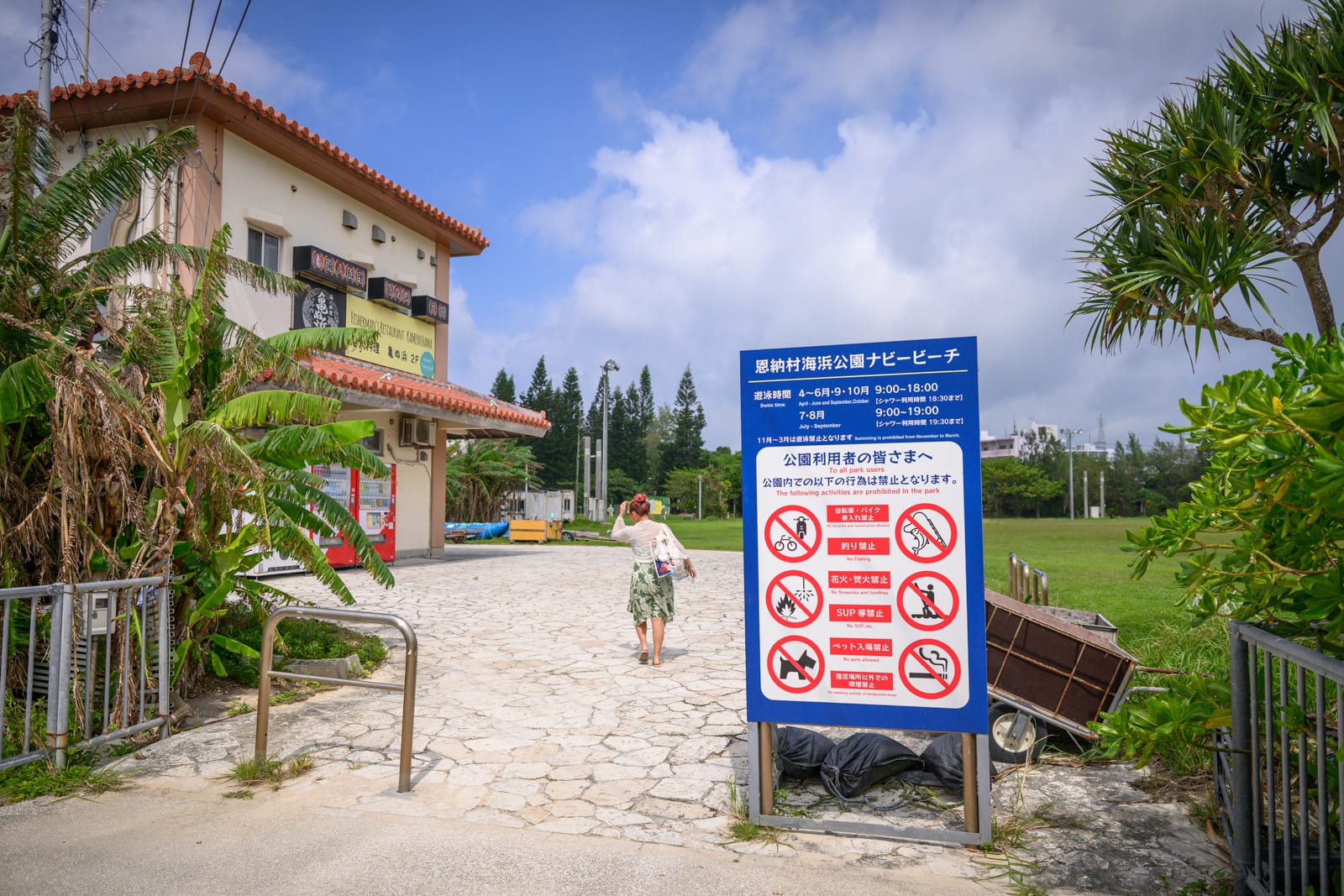 日本沖繩恩納村海濱公園｜街道,建築物,告示牌,綠地,女人,藍天白雲。穿著長裙的女人背對鏡頭行走於石板路上，右側立著藍色告示牌，前方為紅瓦白牆建築與大片草地，周圍點綴著熱帶植物。