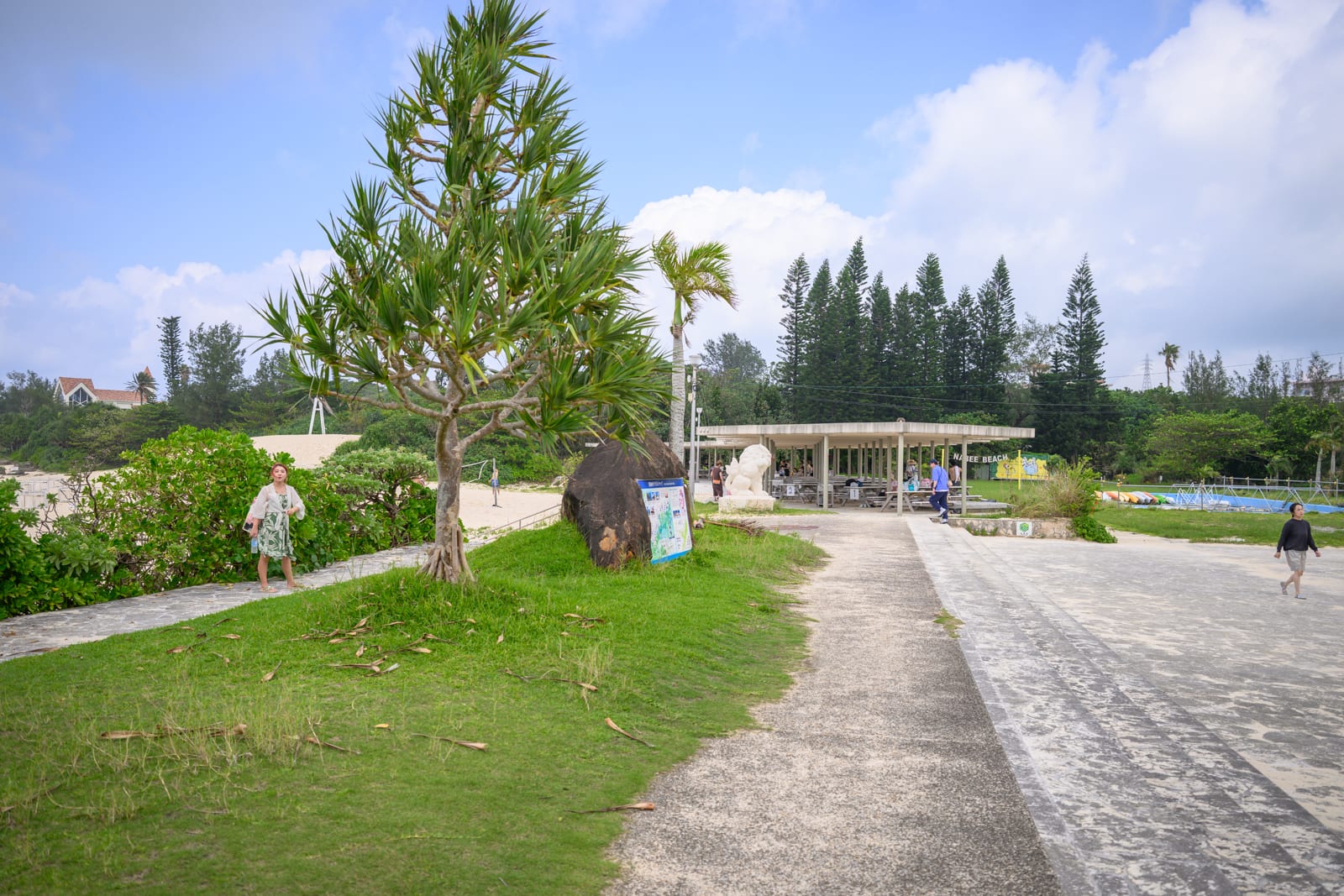 日本沖繩恩納村海濱公園｜樹木、草皮、步道、涼亭、行人、海邊、藍天，戶外空地上鋪設灰白石板路，左側草皮立著帶刺綠葉植物，遠處有白色遮陽棚結構與密集的針葉樹木，天空帶有大片白雲。