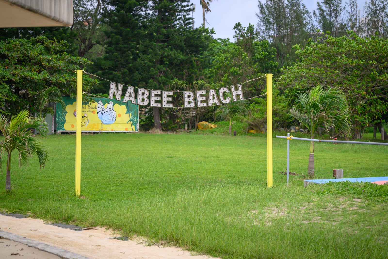 日本沖繩恩納村海濱公園｜草地、立牌、棕櫚樹、招牌、海灘，黃色金屬支架懸掛著「NABEE BEACH」字樣，背景為濃密的綠色樹木與彩繪牆面。