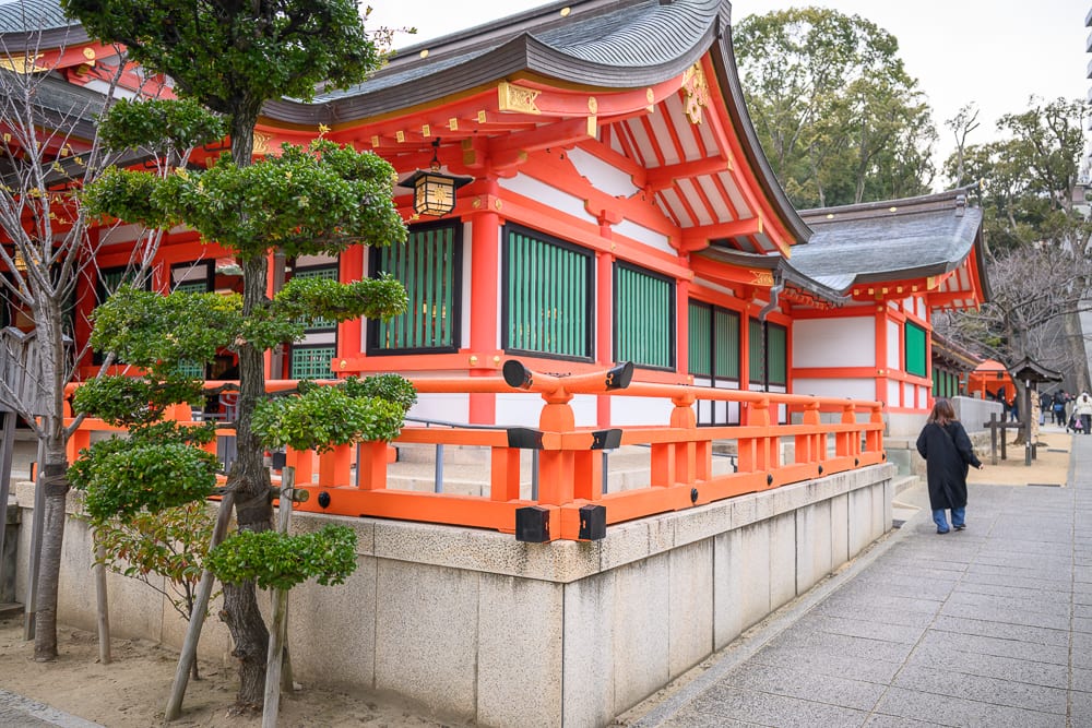 日本神戶-生田神社｜寺廟，橘紅色柱廊，黑瓦屋頂，修剪整齊的松樹，石造圍牆，鋪面步道，穿深色大衣的行人。