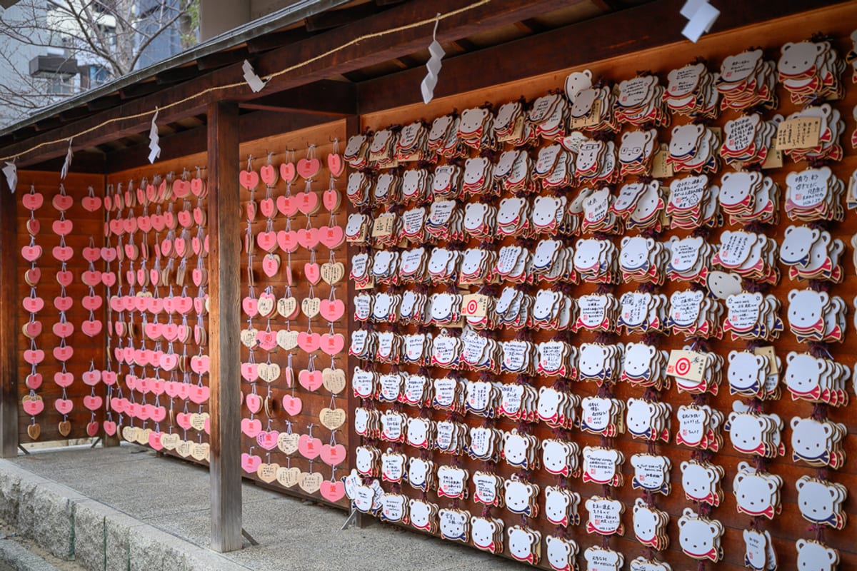 日本神戶-生田神社｜木造神社走廊、祈福木牌、熊形繪馬、心形繪馬、繩索、紙垂、木柱。廊下懸掛大量白色熊形與粉紅心形繪馬，密密麻麻排列在木牆上，邊緣繫著紅色緞帶，上方有橫繩與白色紙垂。