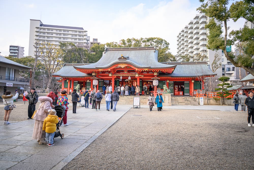 日本神戶-生田神社｜寺廟、廣場、人群、嬰兒車、建築物，灰色石板廣場上聚集著參拜人潮，朱紅色的傳統寺廟建築坐落其中，周邊環繞著現代高樓住宅。