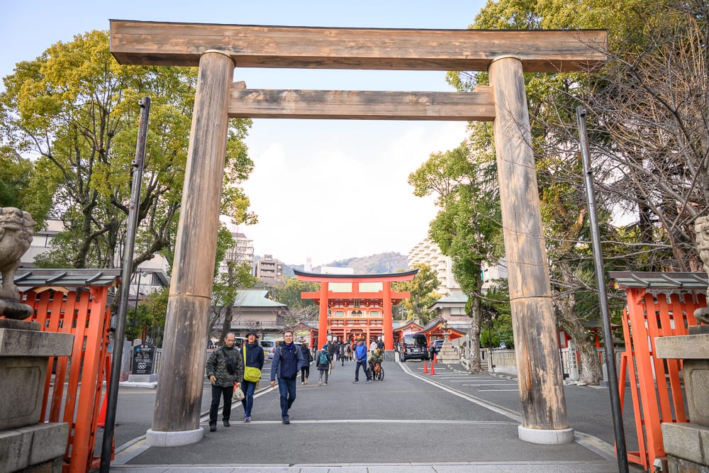 日本神戶-生田神社｜木造鳥居、石板路、紅色牌坊、參拜人群、神社、綠樹。巨大的木造鳥居矗立在道路前方，兩側有紅色柵欄與石燈籠，多名遊客正沿著灰色的石板路向前行走，遠處可見紅色建築與山頭。