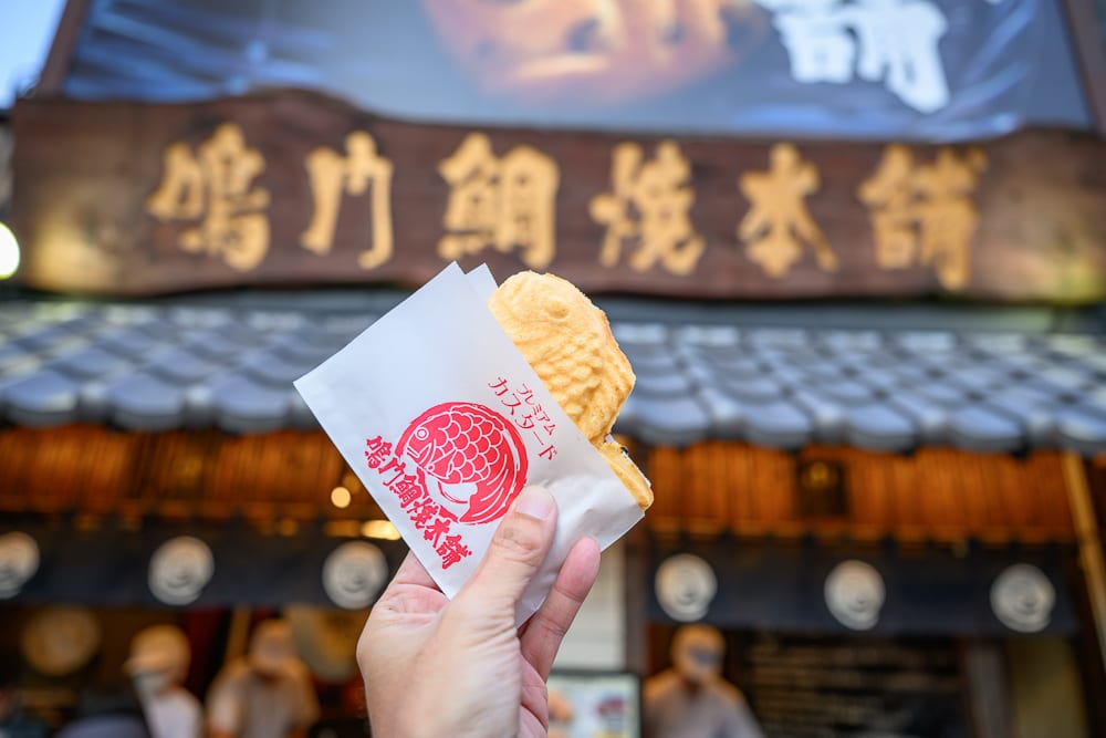 日本神戶-生田神社｜料理，鯛魚燒，紙袋，招牌，屋簷，店面。 手持裝在印有紅色圖案紙袋內的鯛魚燒，背景為懸掛金字木製招牌與灰色瓦片屋簷的日式店舖。
