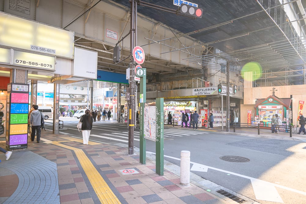 日本神戶-生田神社｜街道，行人，高架橋，招牌，斑馬線，商店。高架鐵路下方交錯著十字路口，行人穿梭於鋪面平整的街道。柱子掛著導覽地圖與標誌，兩側店家招牌林立，遠方車輛停等，灰色柏油路面劃有白色標線。