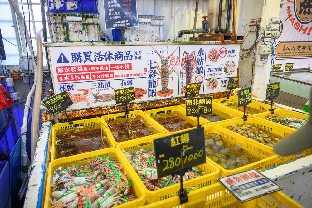 台中阿布潘水產｜市場攤位，黃色塑膠籃，活體海鮮，螃蟹，蝦，貝類，價格標示牌，水產看板，白色牆面。