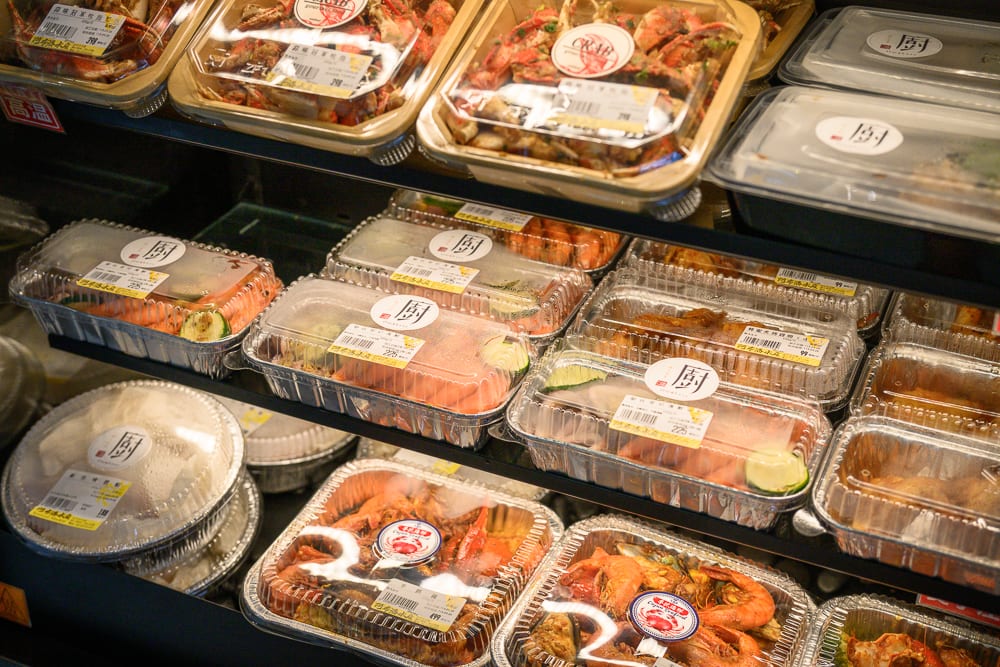 台中阿布潘水產｜超市冷藏櫃內擺放多層塑膠盒裝料理，包含蝦子、螃蟹、炸物與米飯等便當，透明蓋上貼有價格標籤與圓形標誌。