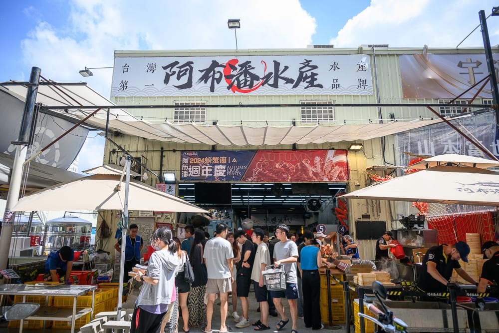 台中阿布潘水產｜招牌、建築、人群、遮陽傘、攤位，水產店門口聚集眾多顧客排隊等候，上方懸掛白色布幔與大型招牌，兩側設有多支米色遮陽傘，現場人員正忙碌地處理業務。