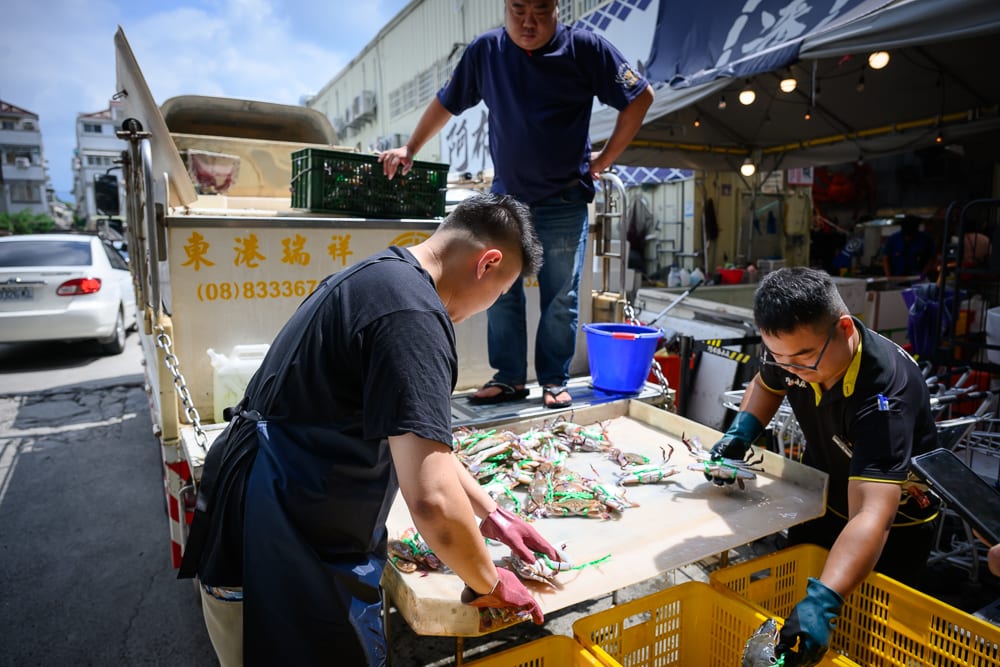台中阿布潘水產｜貨車、攤販、螃蟹、工作人員、街道、塑膠籃，三名男子在戶外貨車旁作業，桌上散落多隻螃蟹，周圍堆疊黃色塑膠籃。