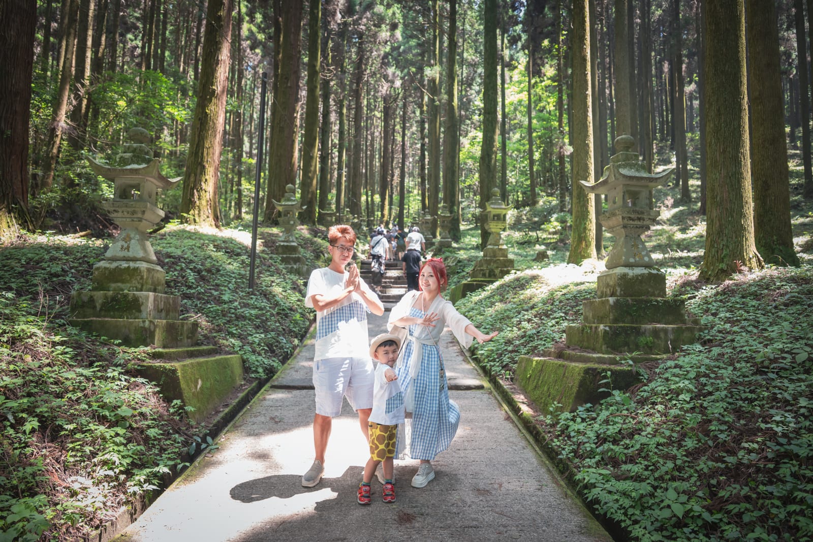 日本熊本-上色見熊野座神社｜森林，步道，石燈籠，三人，家庭，高聳樹木，苔蘚，山徑。一家三口站在森林石階步道中央擺出動作，兩側立有斑駁石燈籠與茂密綠草，背景為筆直參天的針葉林。