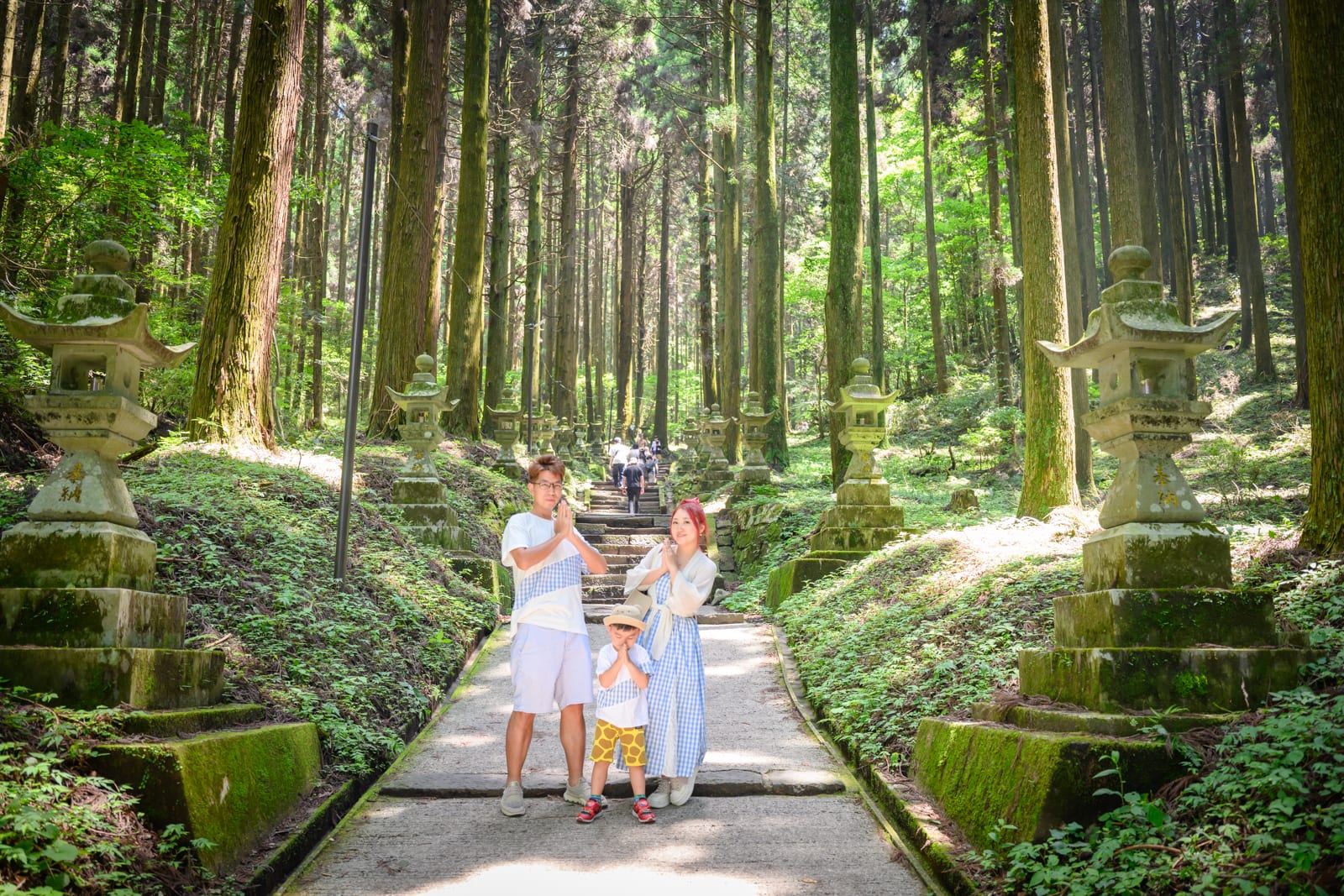 日本熊本-上色見熊野座神社｜三人家庭、步道、石燈籠、高聳樹林、苔蘚石階。一家三口站在鋪面平坦的步道中央，兩側矗立長滿青苔的石燈籠與茂密針葉林，背景延伸長階梯。