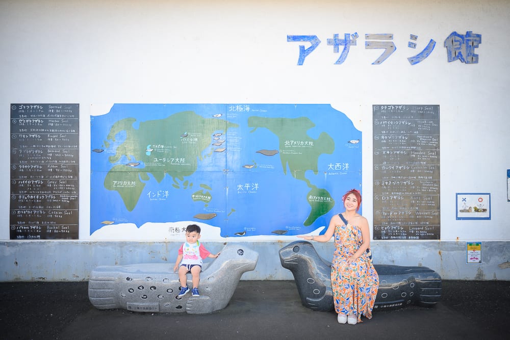 日本小樽-小樽水族館｜婦人、幼童、海豹雕像、白色牆面、世界地圖、日文字標示。婦人身穿印花長裙坐在黑色海豹雕像上，幼童坐在左側灰色海豹雕像上，背後為白色牆面，牆面上懸掛藍色世界地圖與日文字標示牌。