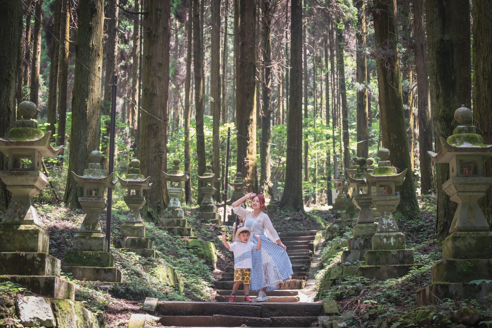日本熊本-上色見熊野座神社｜林道、石燈籠、石階、母子、樹木，高聳的針葉林中鋪設石階，兩側排列著青苔斑駁的石燈籠，一名穿藍白長裙的女子與小男孩站在中央石階上，林間光影錯落。