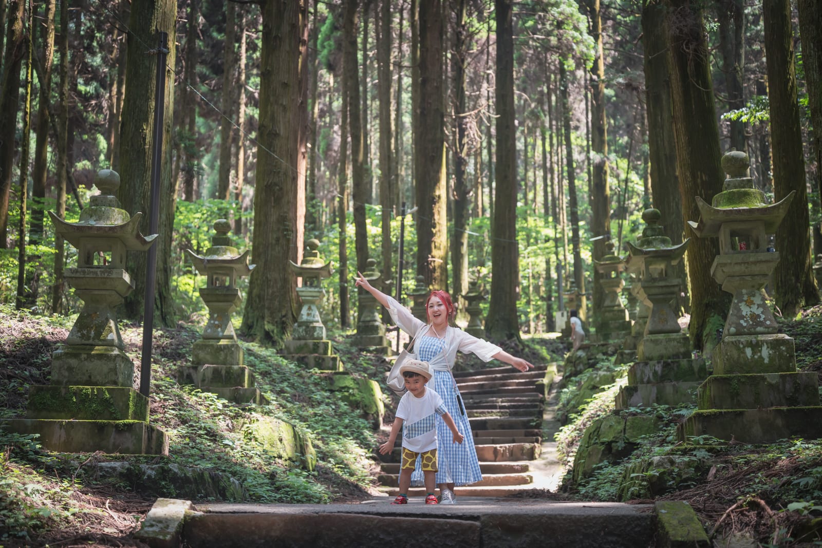 日本熊本-上色見熊野座神社｜林道、石燈籠、石階、母子、樹木，高聳筆直的針葉林夾道，佈滿青苔的石燈籠排列在步道兩側。母親雙臂張開，幼童戴著圓帽站在前方，背景為綿延向上的石階與茂密森林。