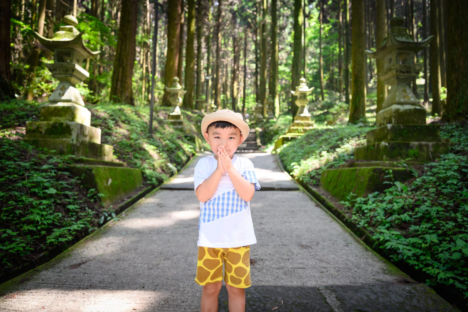 日本熊本-上色見熊野座神社｜男孩，石燈籠，林道，樹林，苔蘚，石階，短褲，帽子。小男孩戴著帽子與雙手合十，站在被林木與苔蘚石燈籠夾夾的石板路上，背景延伸著筆直樹幹。