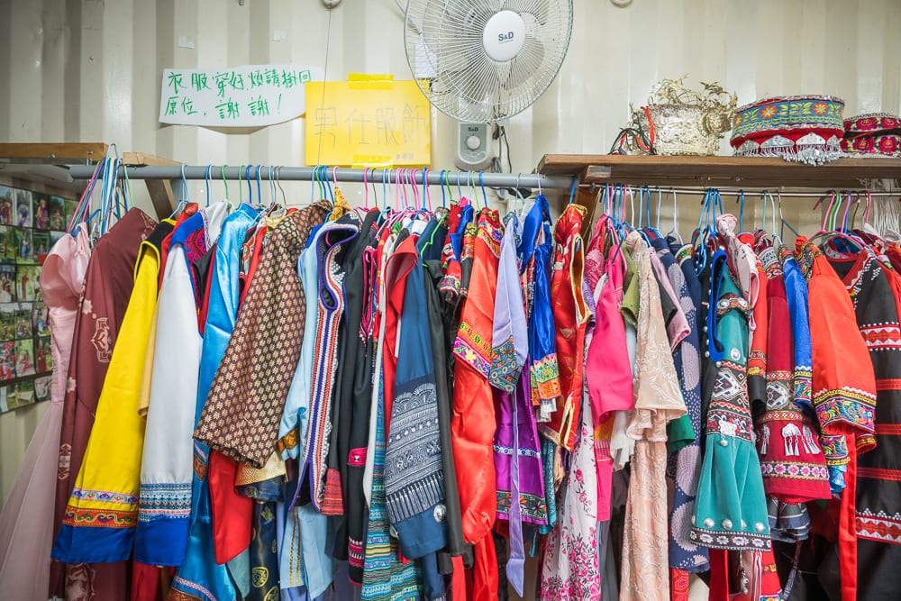 桃園玉飲翡翠文化城完整指南|庭園散步、玉石與民族風陳列 10 桃園玉飲翡翠文化城|室內,衣架,傳統服飾,電風扇,告示牌。牆面懸掛金屬掛衣桿,整齊排列多件色彩鮮艷的民族風服飾與衣物,上方設有置物架,擺放著帽飾。