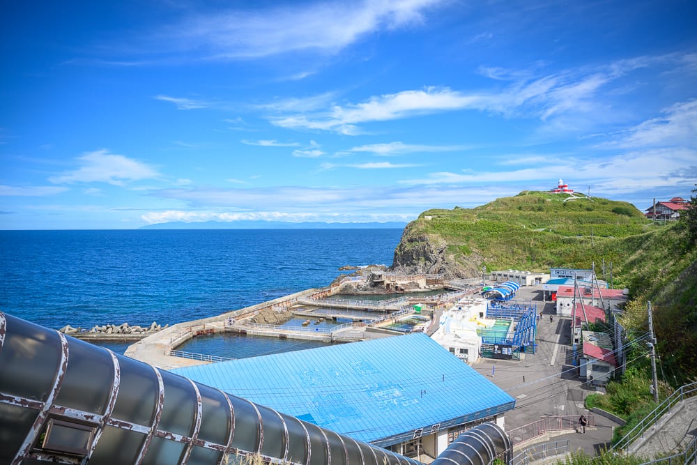 日本小樽-小樽水族館｜藍天、大海、海岸岬角、藍色屋頂建築、海水浴場、山丘、燈塔。蔚藍天穹下廣闊海景，沿岸設有藍頂房舍與階梯水池設施，遠方高處聳立小型燈塔，前方為深色拱形屋頂結構。