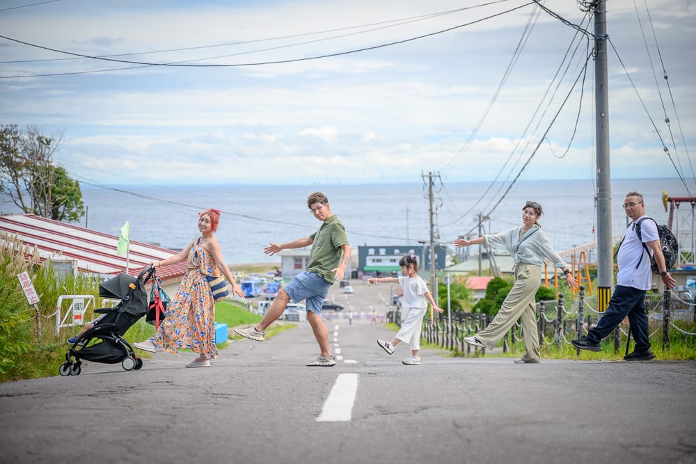 日本小樽-小樽水族館｜街道、海邊、電線杆、推車、行人，五個人在坡道上排成一列單腳站立，背景為蔚藍海水與陰暗天空，道路兩旁設有護欄與房舍。