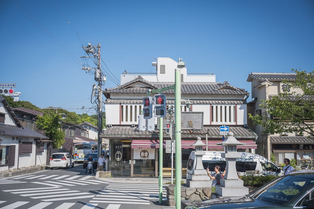 日本鶴岡八幡宮完整指南|參道散步、石階與周邊街區 7 日本鎌倉-八幡宮|街道,斑馬線,傳統建築,電線桿,汽車,行人,紅綠燈,石燈籠。晴朗藍天下的十字路口,白色日式瓦房與店鋪林立,車輛停等紅燈,行人站在路口,電線桿交錯。