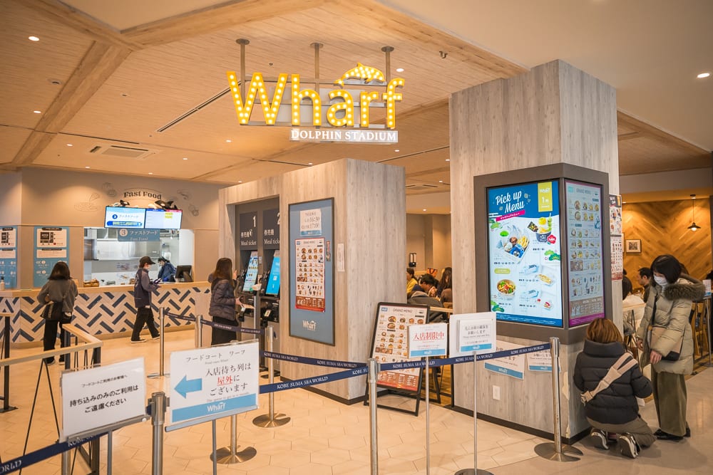 日本神戶-須磨海洋世界｜室內、餐廳、點餐機、招牌、排隊告示、人群。Wharf燈光招牌懸掛於木紋天花板下，下方設有多台直立式觸控點餐機與藍色排隊護欄，數位螢幕顯示菜單，數位看板亮起，顧客站立操作或在櫃檯前走動。