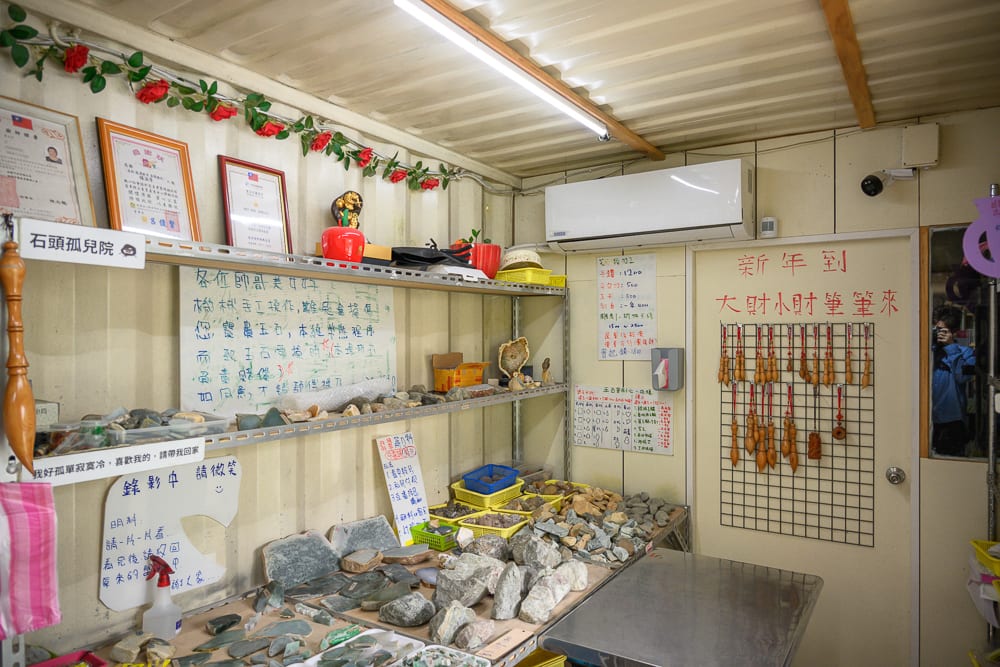 桃園玉飲翡翠文化城完整指南|庭園散步、玉石與民族風陳列 5 桃園玉飲翡翠文化城|室內,層架,原石,告示牌,門,招牌。鐵皮屋內設有多層金屬架,擺放大量大小不一的灰色原石與玉石,牆面貼有手寫說明、獎狀與紅色春聯,日光燈管照明。