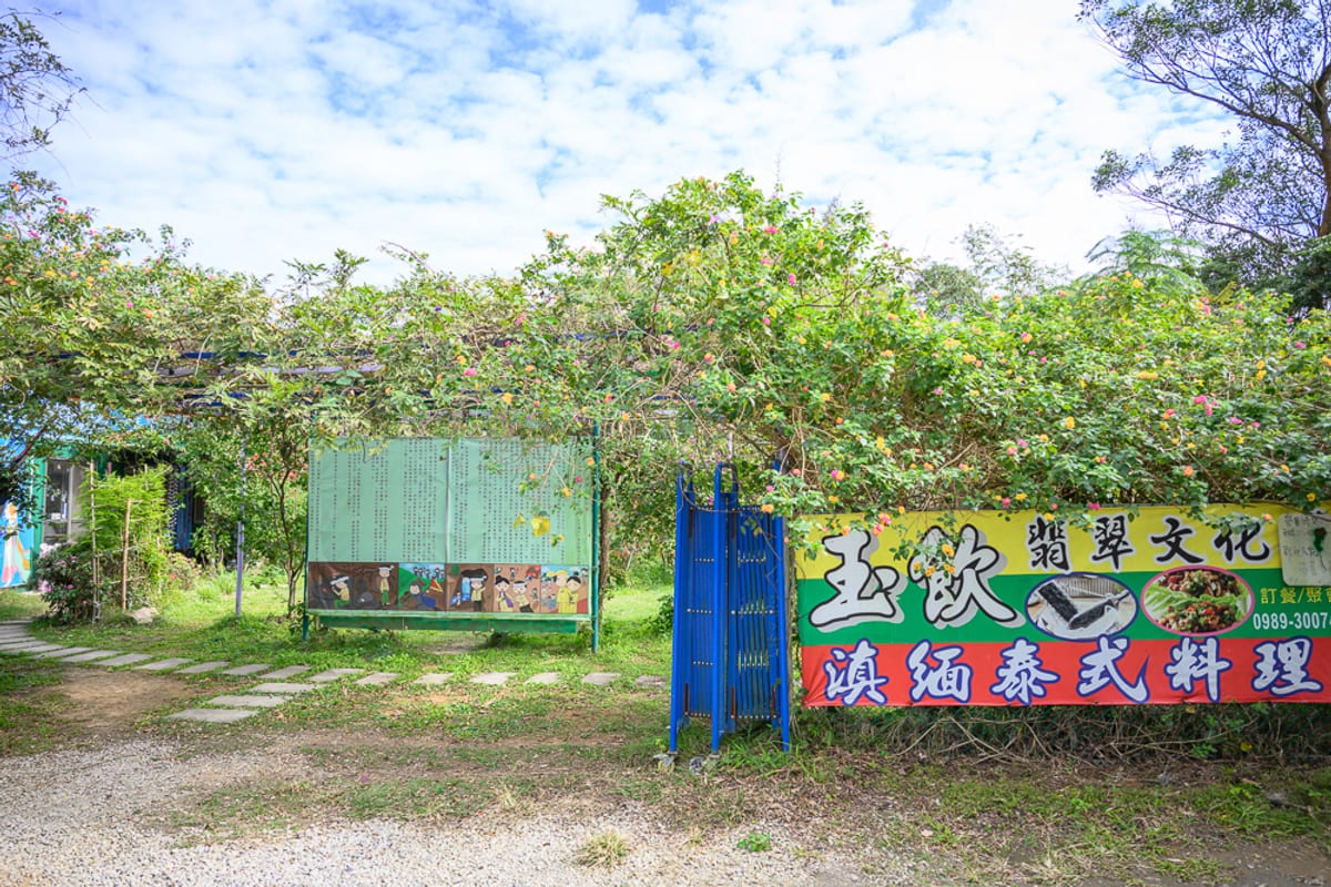 桃園玉飲翡翠文化城完整指南|庭園散步、玉石與民族風陳列 1 桃園玉飲翡翠文化城|戶外、草地、招牌、樹木、藍天,玉飲翡翠文化滇緬泰式料理的彩色布條懸掛在枝葉繁茂的藤架旁,前方設有藍色伸縮門與綠色圖文看板,地面鋪有石板步道。