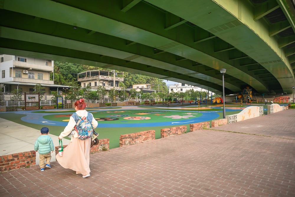 苗栗滑步車公園｜綠色高架橋、磚牆街道、彩色遊樂場、背背包的女性與幼童，遠處有低矮房舍與山丘，地面鋪設紅磚與彩色地墊。