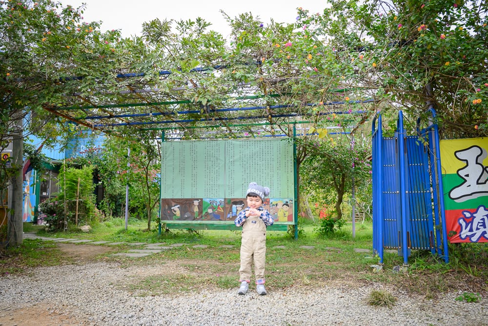 桃園玉飲翡翠文化城完整指南|庭園散步、玉石與民族風陳列 3 桃園玉飲翡翠文化城|小孩、綠地、藤架、告示牌、藍色柵欄,戶外庭園環境,碎石地面鋪著石板步道,背景有濃密枝葉覆蓋的棚架與建築結構。