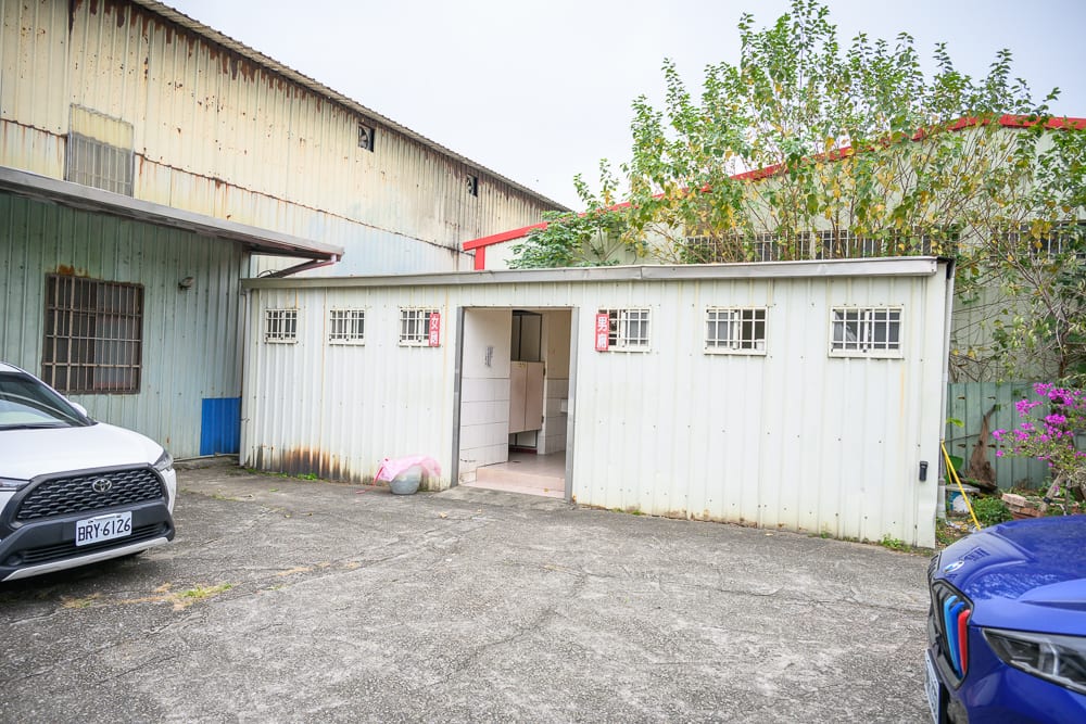 桃園白蛇廟｜戶外空地，白色鐵皮屋，建築物，汽車，樹木，窗戶，牆面。地坪鋪設灰色水泥，前方兩側停放白色與藍色車輛，後方為黃色鐵皮廠房，牆上有紅色字體標示。