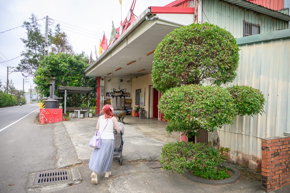 桃園白蛇廟｜街道、寺廟、樹木、推車、人。白衣長裙女性推著嬰兒車，走在柏油路邊的磚牆建築旁，前方為廟宇入口，右側立著修剪圓球狀的景觀樹，左側有電線桿與道路。