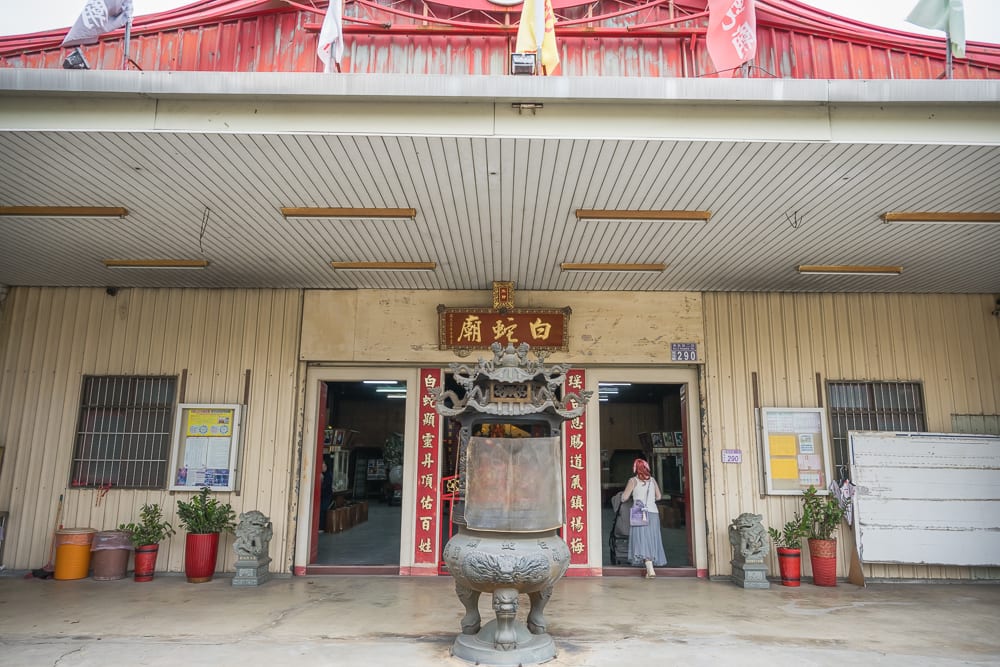 桃園白蛇廟｜廟宇，香爐，石獅，招牌，門聯，盆栽，鐵皮屋頂，人物。正面拍攝白蛇廟外觀，建築採用鐵皮與金屬結構，門口放置大型石雕香爐與石獅，兩側有盆栽與窗戶，紅字招牌與門聯懸掛於入口上方，一名女子正走入室內。