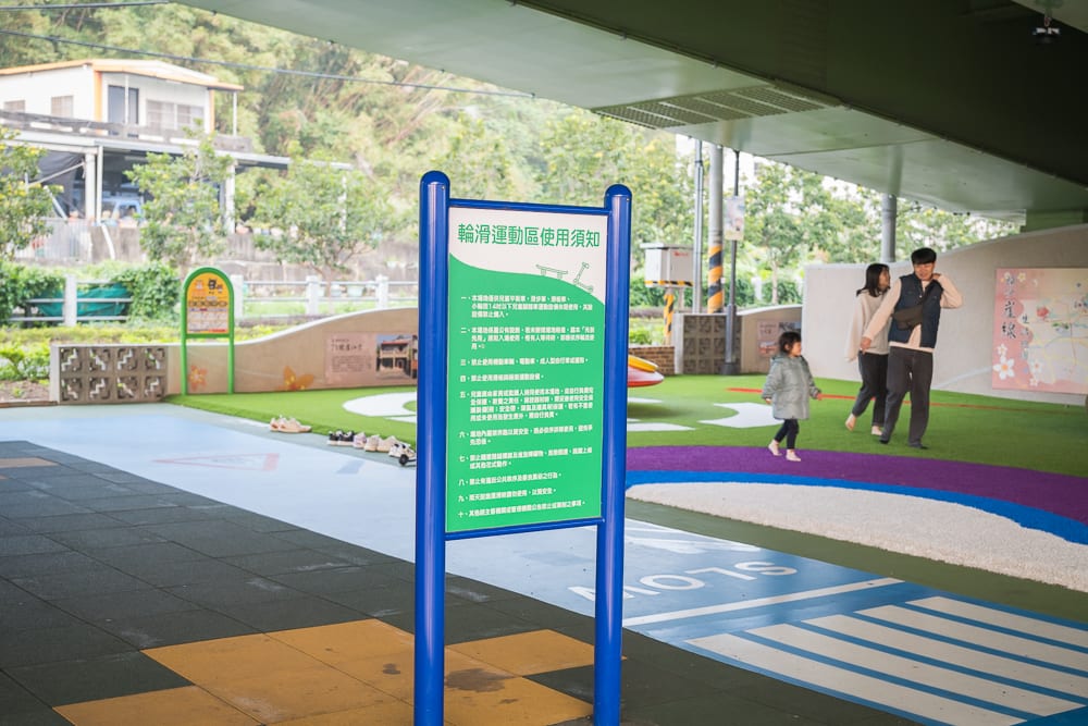 苗栗滑步車公園｜高架橋下，告示牌，溜冰場，人工草皮，行人，孩童，灰色水泥地柱，藍色鐵架，房屋，綠樹。
