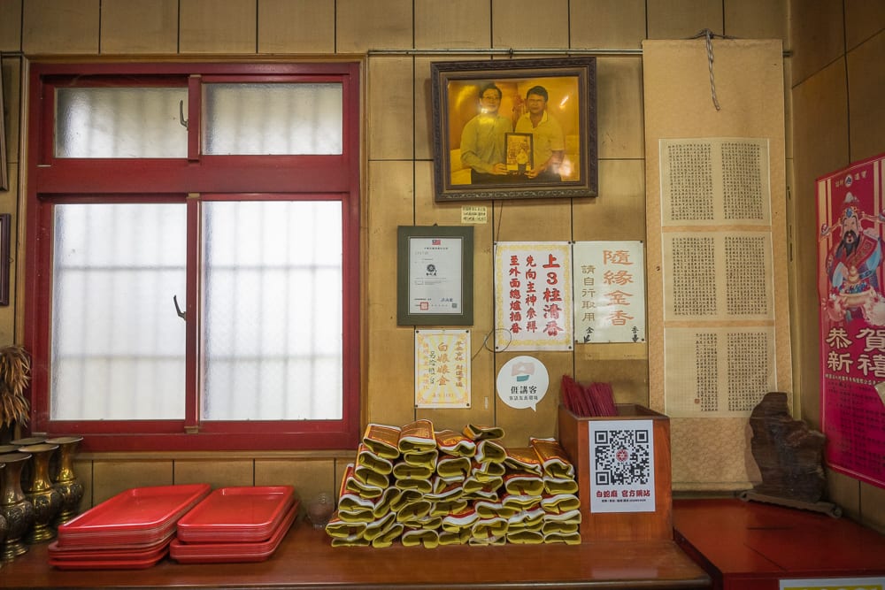 桃園白蛇廟｜室內，木牆，紅框窗戶，金紙，托盤，相框，公告紙張，壁掛捲軸。木桌上堆疊黃色金紙與紅色托盤，牆面掛著鏡框照片、參拜說明告示與書法捲軸，透出日光。