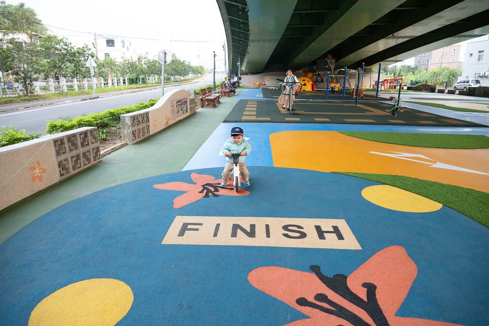 苗栗滑步車公園｜高架橋下空間，幼童，滑步車，彩色跑道，花朵圖案，終點線標示，遊樂場，遠方道路，建築物。